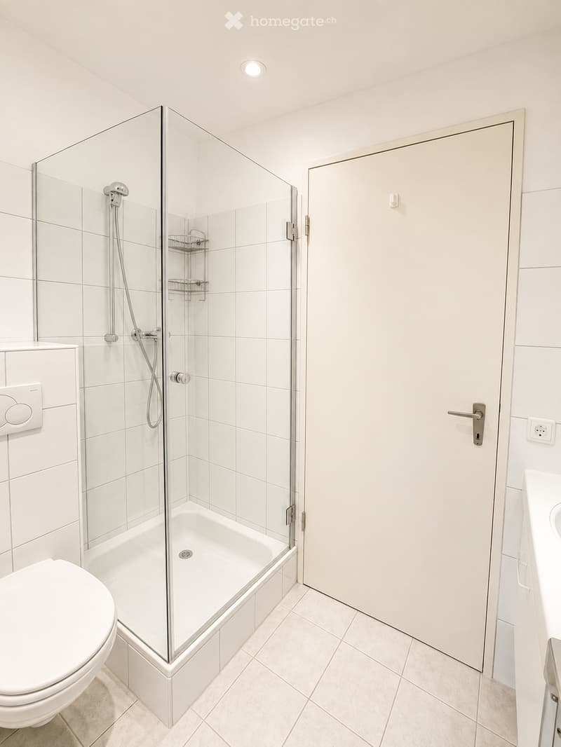 Modern bathroom with white tiles, a bathtub enclosed by a glass shower enclosure, a toilet, and a sink.