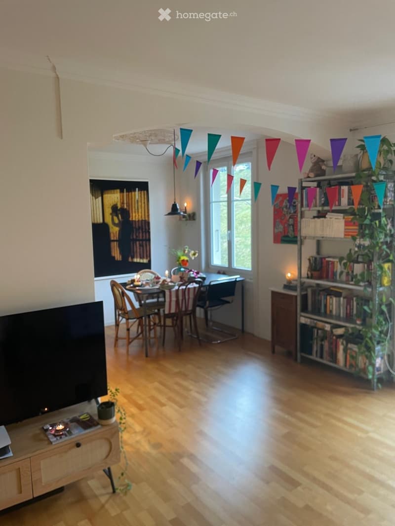 A well-lit living and dining area with wooden flooring, a large TV on a cabinet, a dining table with chairs, and a bookshelf filled with books and decorative items.