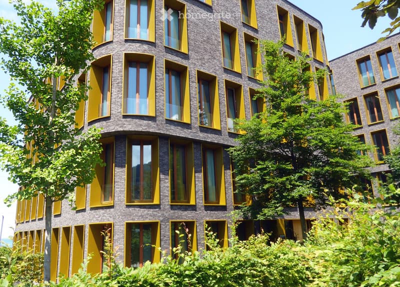 Modern multi-story building with dark brick facade and bright yellow window frames, surrounded by lush greenery.