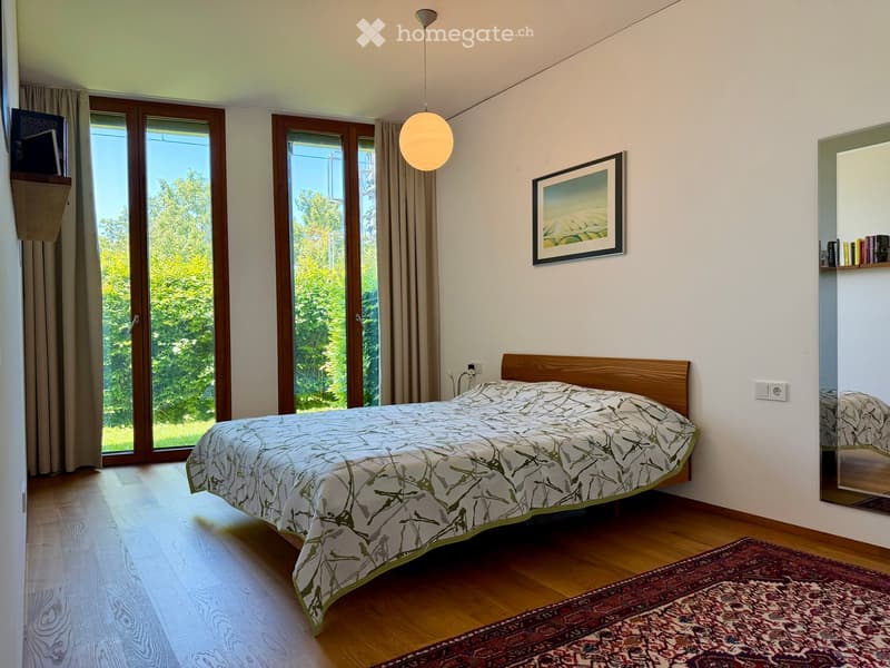 Bright bedroom with wooden flooring, large windows with curtains, a bed with patterned bedding, a mirror, and a picture frame on the wall.