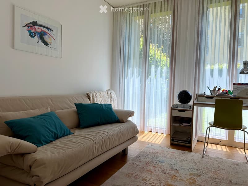 A cozy living room with a beige sofa, teal throw pillows, a small wooden side table with a globe, a desk with a chair, and a large window with sheer curtains.