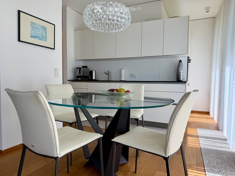 Modern kitchen with white cabinets, a glass dining table surrounded by white chairs, a fruit bowl, and a coffee maker on the counter.