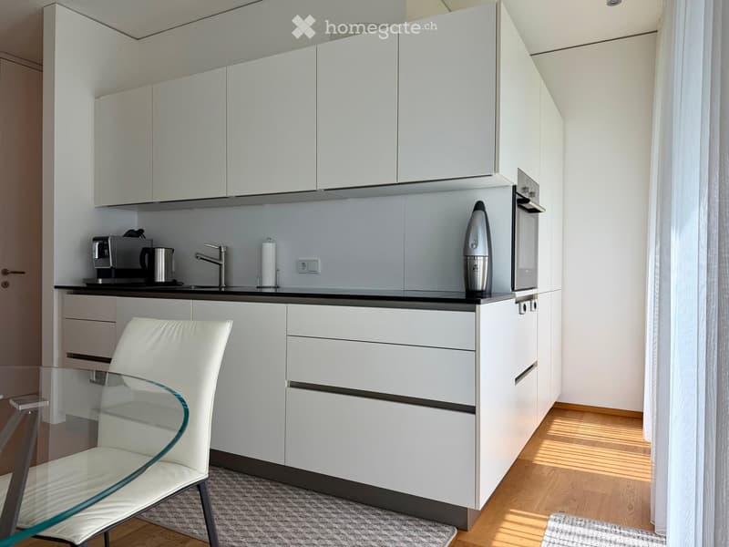 Modern kitchen with white cabinets, black countertop, a white chair, a coffee maker, and an oven.