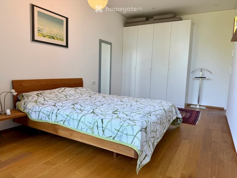 Modern bedroom with wooden flooring, a large bed with a patterned duvet, a nightstand with a lamp, a white wardrobe, a mirror, and a clothing rack.