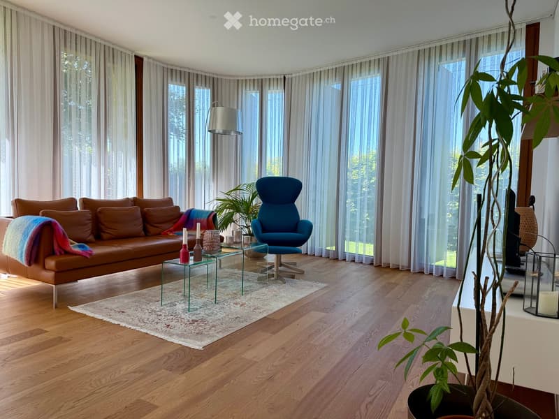 Modern living room with wooden flooring, large windows with sheer curtains, a brown leather sofa, a blue armchair, a glass coffee table, and decorative plants.