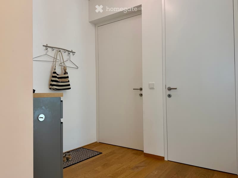 A narrow hallway with light-colored walls, wooden flooring, and two white doors. A small wooden shelf holds a striped bag and a coat hanger, while a small rug lies on the floor near the shelf.