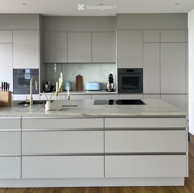 Modern kitchen with white and grey cabinets, marble countertop, built-in oven, sink with gold faucet, and various appliances and decorative items on the counter.