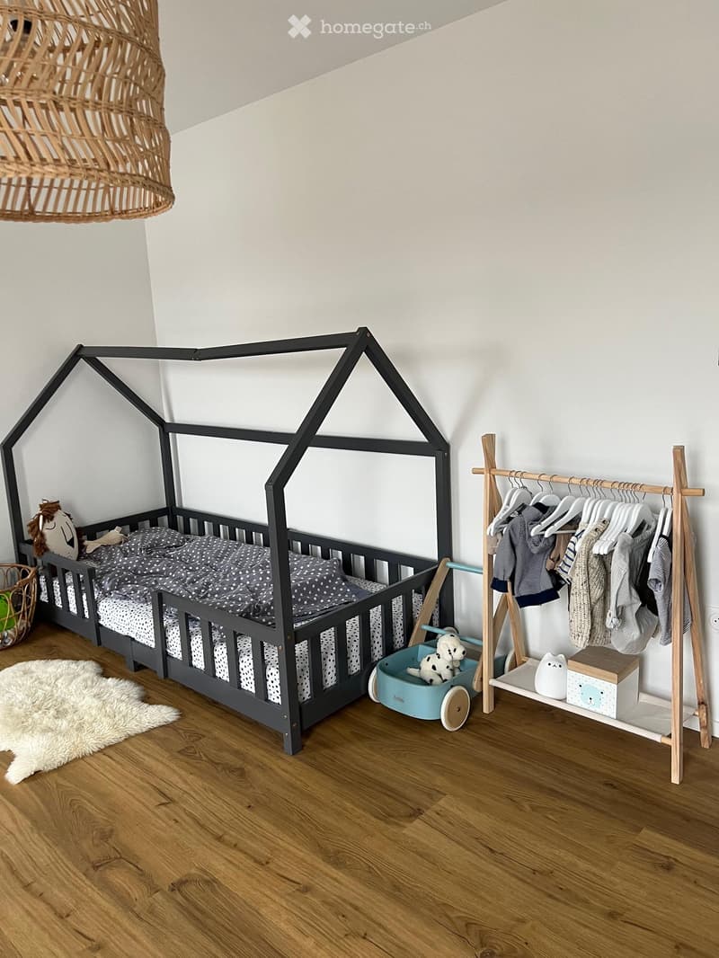 A modern children's bedroom with a geometric bed frame, a hanging woven lamp, a wooden wardrobe, a plush rug, and a small toy cart.