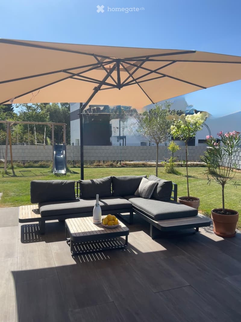 Outdoor patio with a grey sectional sofa, coffee table, umbrella, bottle, and bowl of fruit. The area is surrounded by potted plants and a grassy lawn.