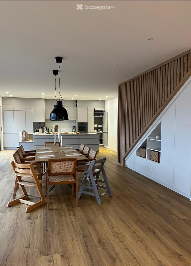 Modern dining area with a wooden dining table, six chairs, and a high chair, connected to a sleek kitchen with white cabinets and stainless steel appliances.