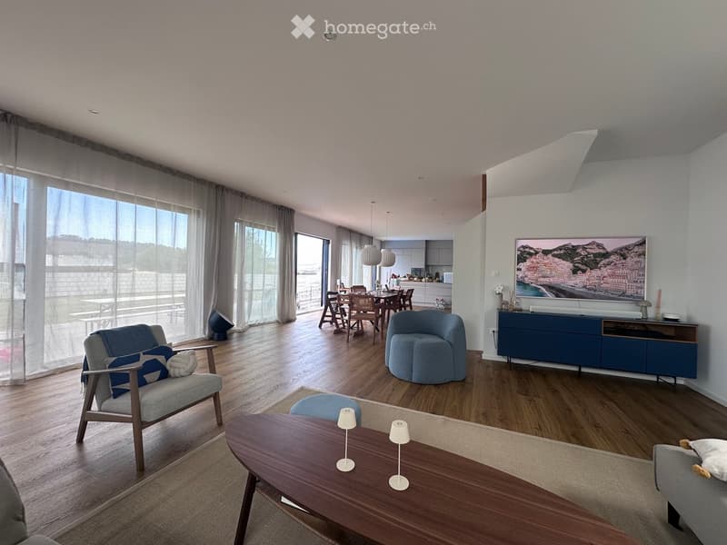 Modern living room with wooden flooring, large windows, and a view. Features include a grey sofa, wooden coffee table, blue accent chair, dining area with a table and chairs, and a blue cabinet with a TV.
