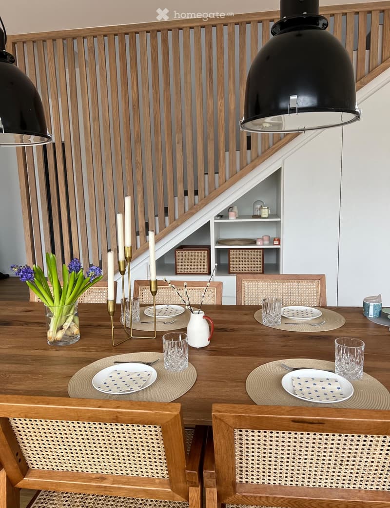 Dining room with a wooden table set for six, surrounded by wicker chairs, with a vase of flowers and candles on the table, and a staircase in the background.
