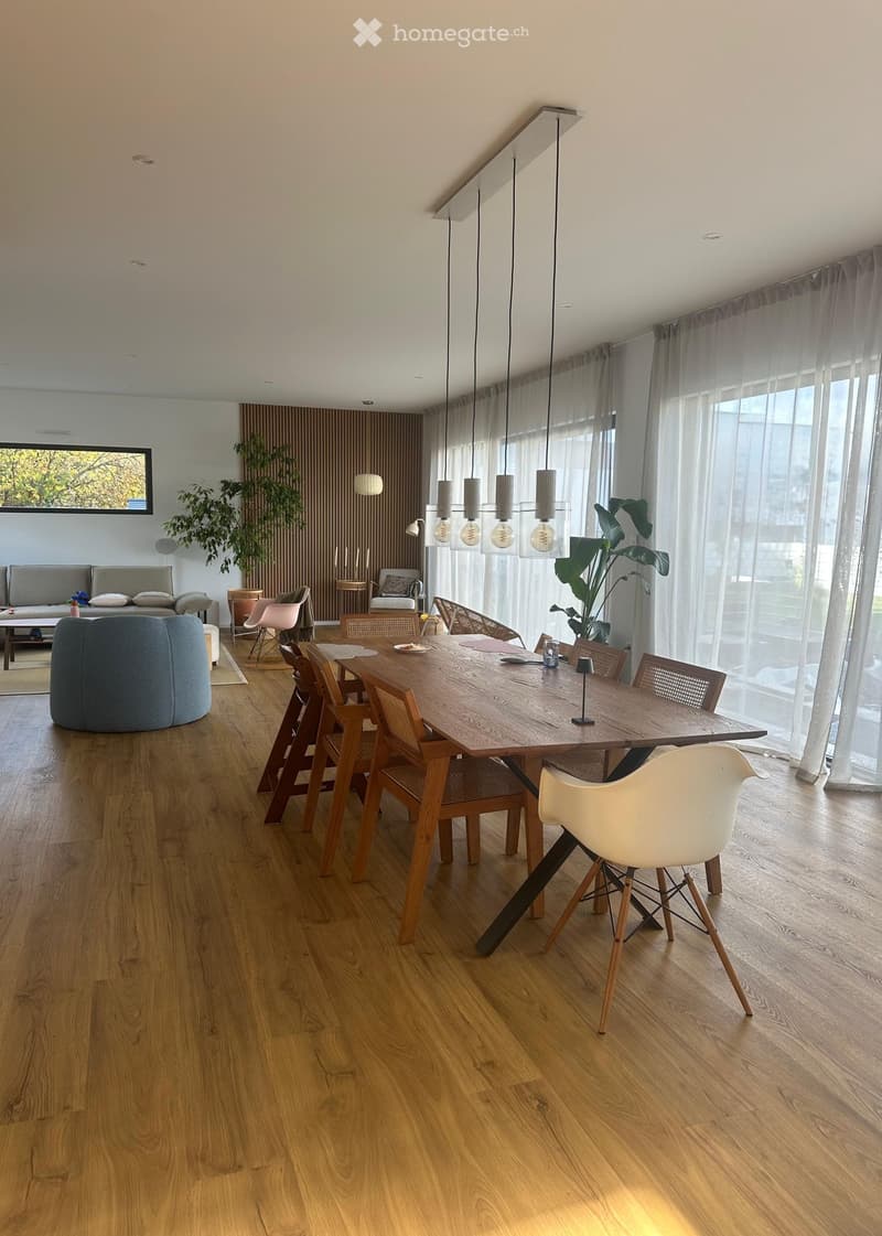 Open-plan living and dining area with wooden flooring, large windows, pendant lights, dining table with chairs, sofa, ottoman, and plants.