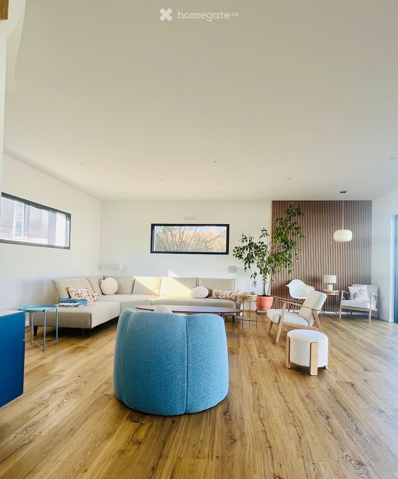 Modern living room with light-colored wooden flooring, a large sectional sofa, a coffee table, several chairs, a potted plant, and decorative lamps.