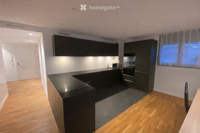 Modern kitchen with dark cabinets, a black countertop, and built-in appliances including a microwave and oven. The space features wooden flooring and a window with blinds.