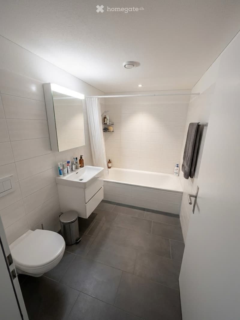 Modern bathroom with white tiles, a sink with a mirror, a toilet, a bathtub, and a shower curtain. A towel hangs on the wall, and a trash can is placed near the sink.