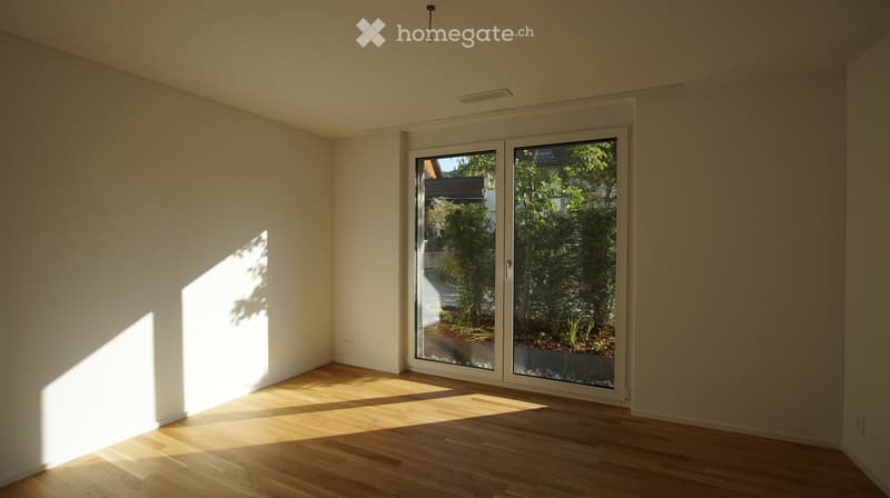 Empty room with light-colored walls and wooden flooring, featuring large glass sliding doors that open to a garden area.