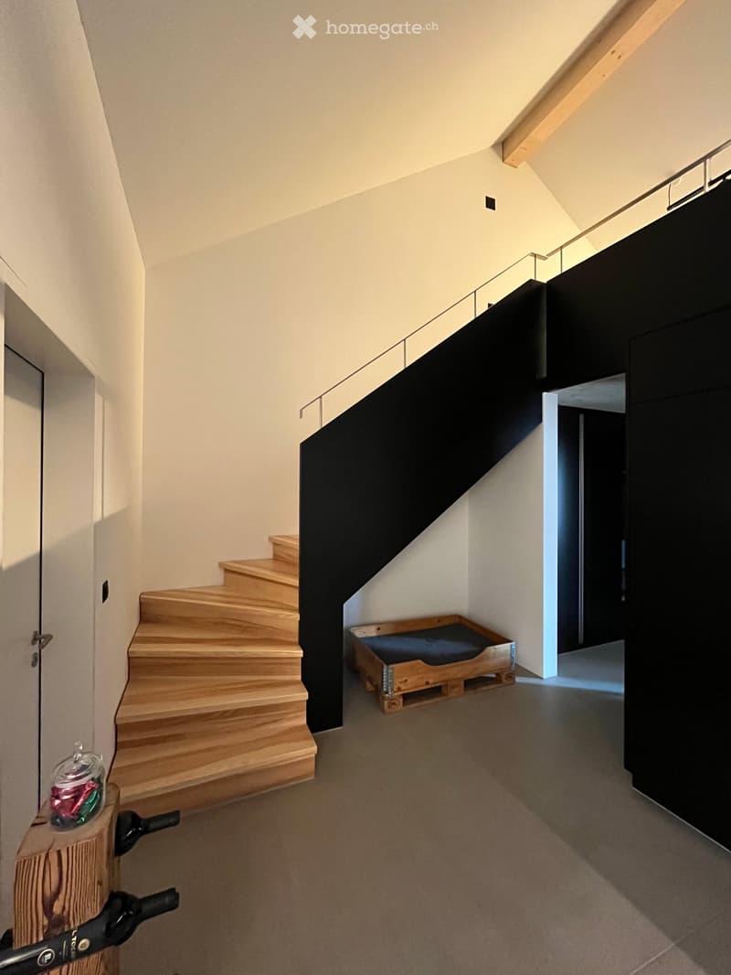 Modern hallway with wooden staircase, sleek black railing, and a small pet bed in the corner.