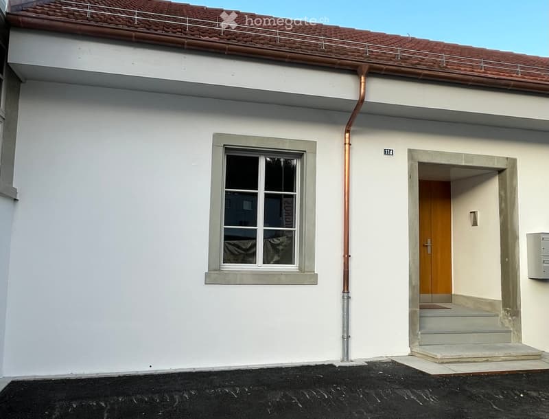 Two-story building with white exterior, red-tiled roof, and a small balcony on the roof. Features a window with a grey frame and a wooden door with a small step leading up to it.
