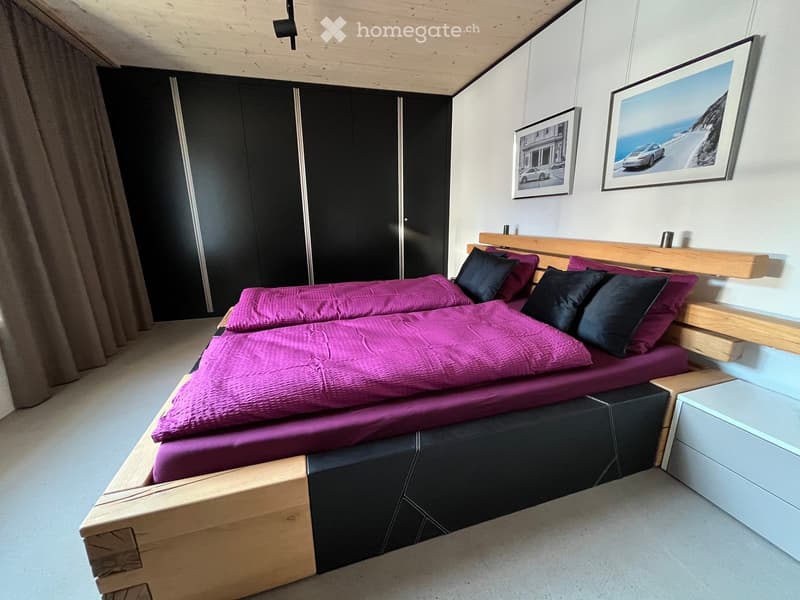 Modern bedroom with a wooden bed frame, purple bedding, black pillows, and a nightstand. Two framed pictures on the wall and a large black wardrobe.