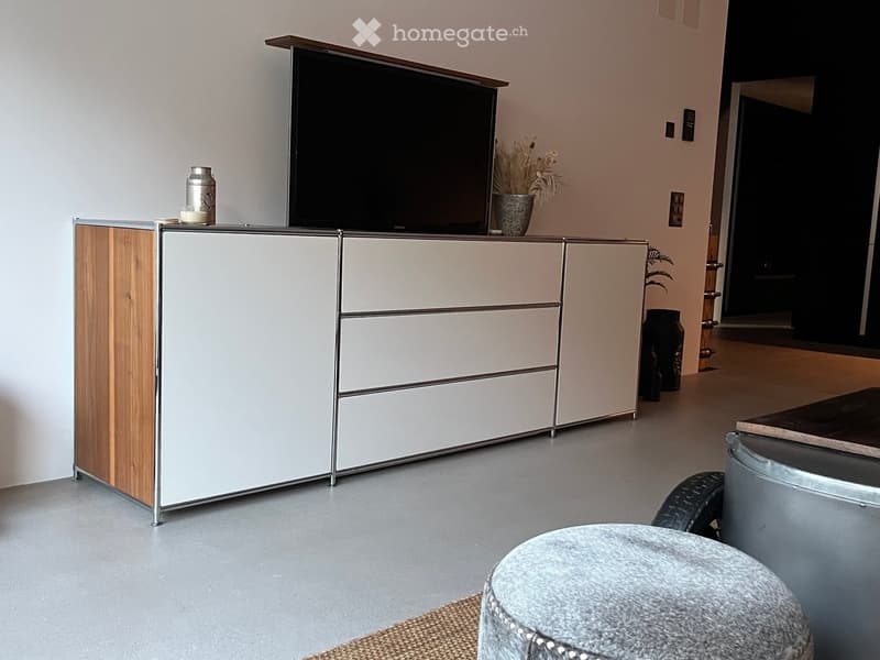 Modern living room with a grey ottoman, a sleek white and wood TV stand, a flat-screen TV, and decorative vases on the stand.