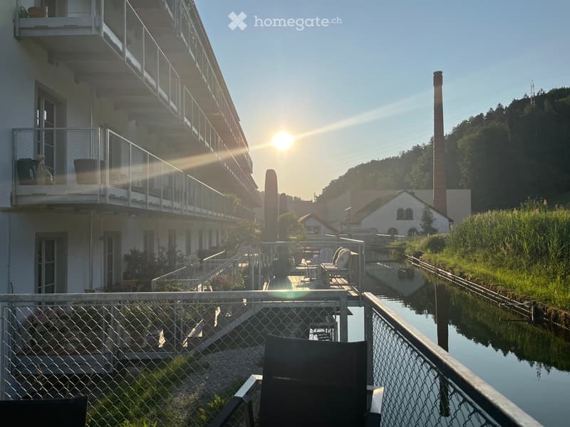 Balcony overlooking a canal with a view of the setting sun, featuring a metal fence, chairs, and a table.