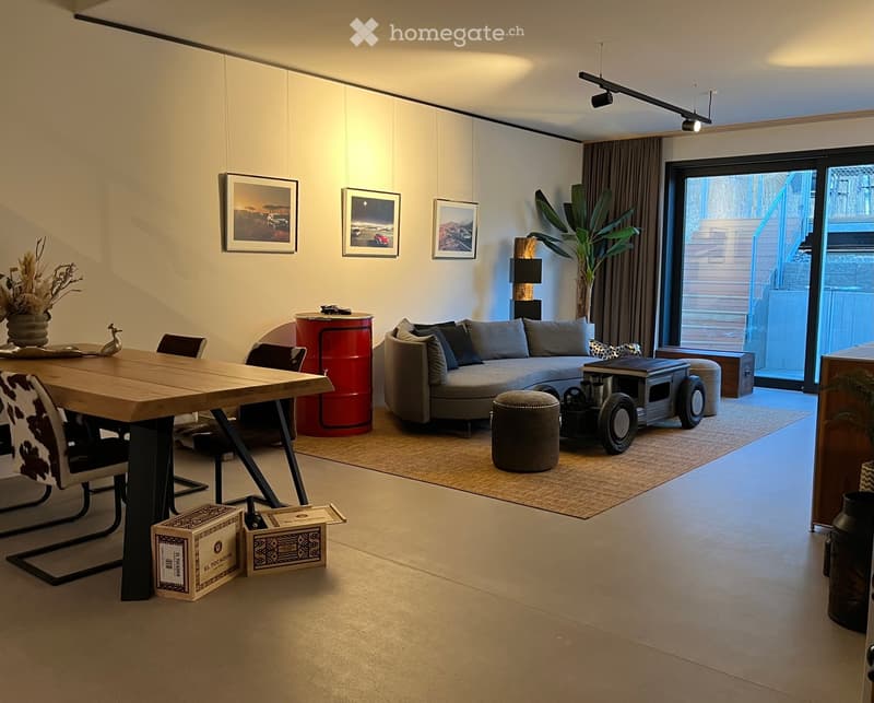 Modern living room with a grey sectional sofa, coffee table, dining table, chairs, and decorative items including a vase and picture frames. The space features large windows with curtains and a sleek, contemporary design.