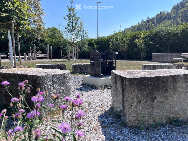 Outdoor area with stone seating blocks, a small metal structure, wooden benches, and playground equipment in the background.