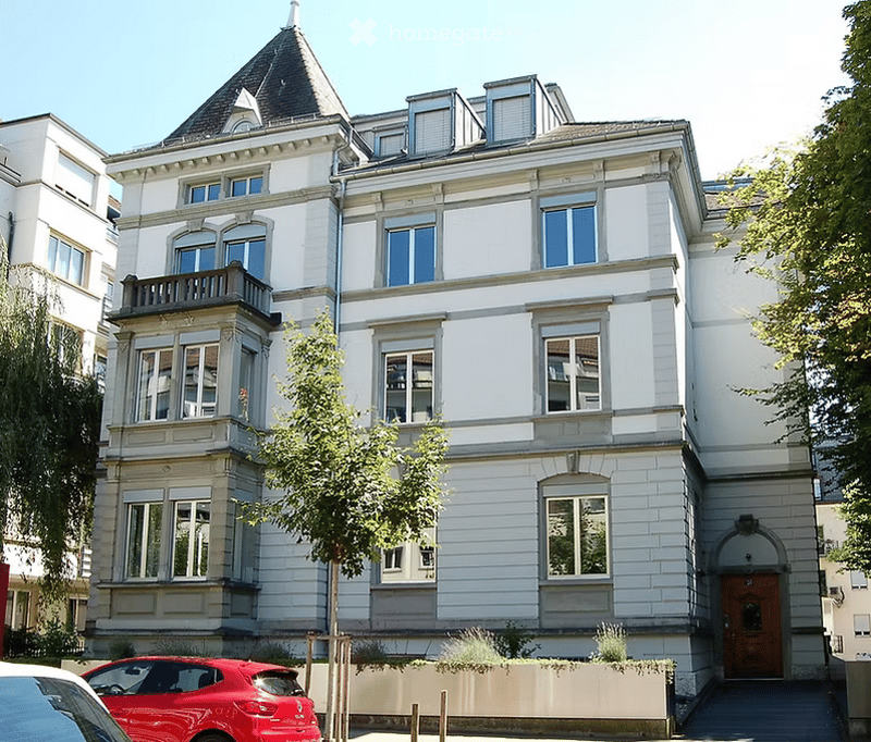 3-story building, white stucco, many windows, gable roof, entrance, balcony