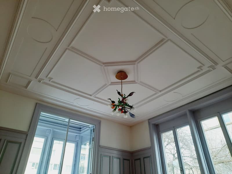 Decorative ceiling with panels and a hanging chandelier. Large windows. Wooden wall panels. Exposed white walls.