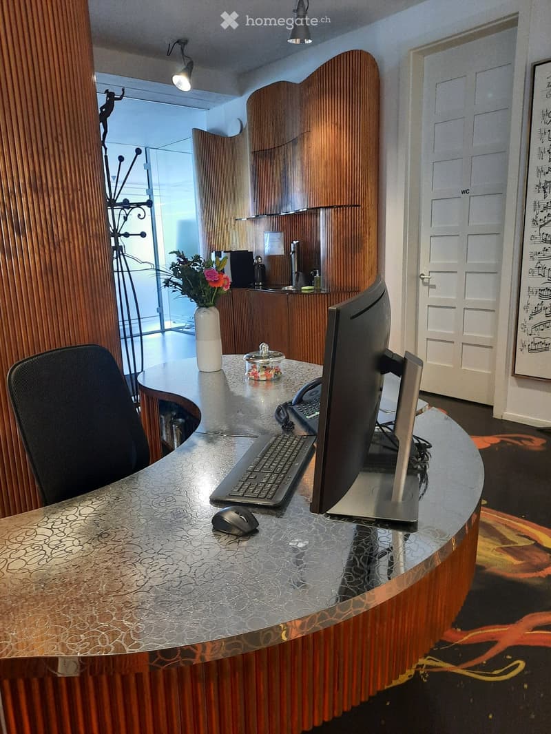 Reception area with a curved wooden desk, computer setup, chair, vase with flowers, and decorative jar.