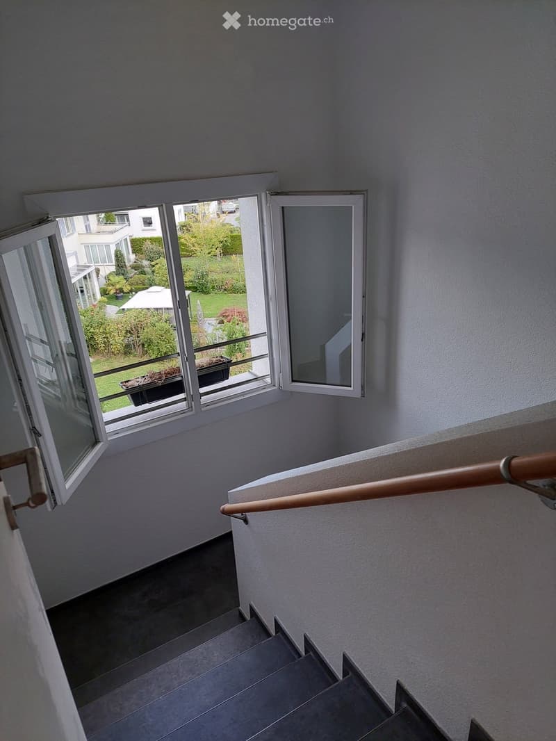 Staircase with wooden handrail, grey steps, and large windows overlooking a garden area with greenery and a patio umbrella.