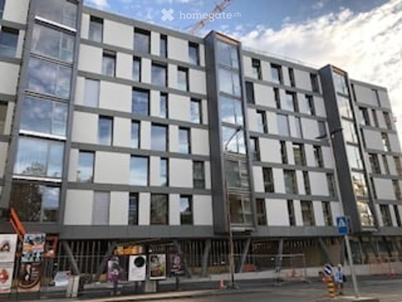 Modern multi-story apartment building with large windows and grey facade, under construction with visible scaffolding and cranes.
