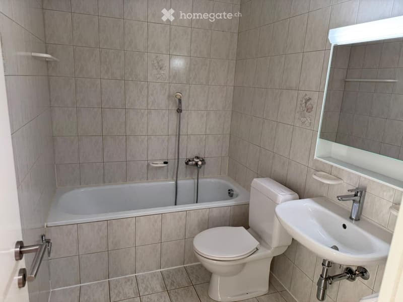 Modern bathroom with a bathtub, toilet, sink, and mirror. The walls are tiled with a light gray pattern, and the floor is tiled in a matching design.