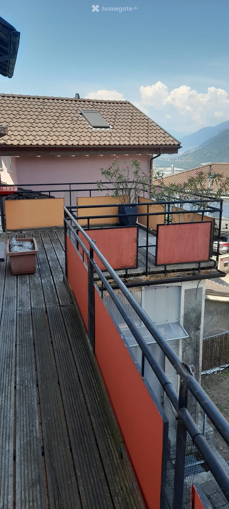 Wooden balcony with metal railing, overlooking a tiled roof and distant mountains, with potted plants and a view of the surrounding area.