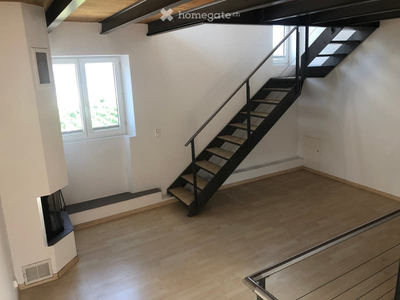 Empty room with wooden flooring, white walls, and a staircase leading upwards. The room has a window and a small landing area.