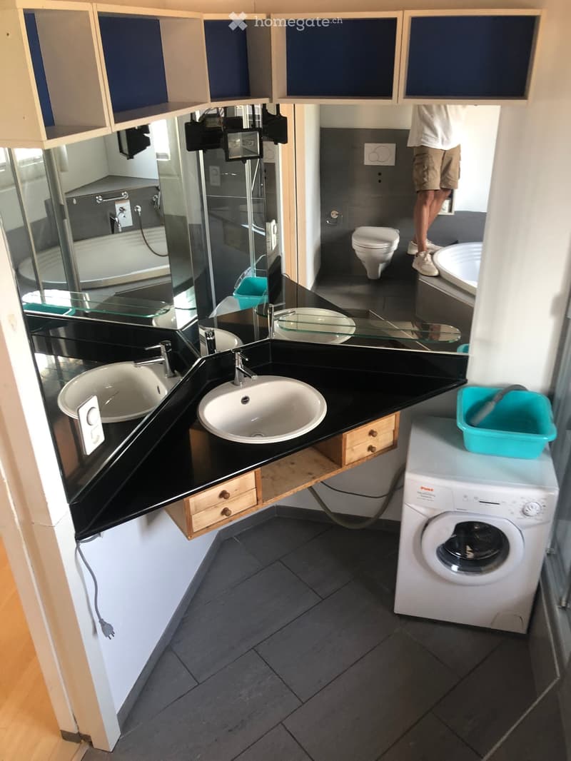 Modern bathroom with double sink, toilet, washing machine, and large mirror. Features dark countertops and tiled floor.