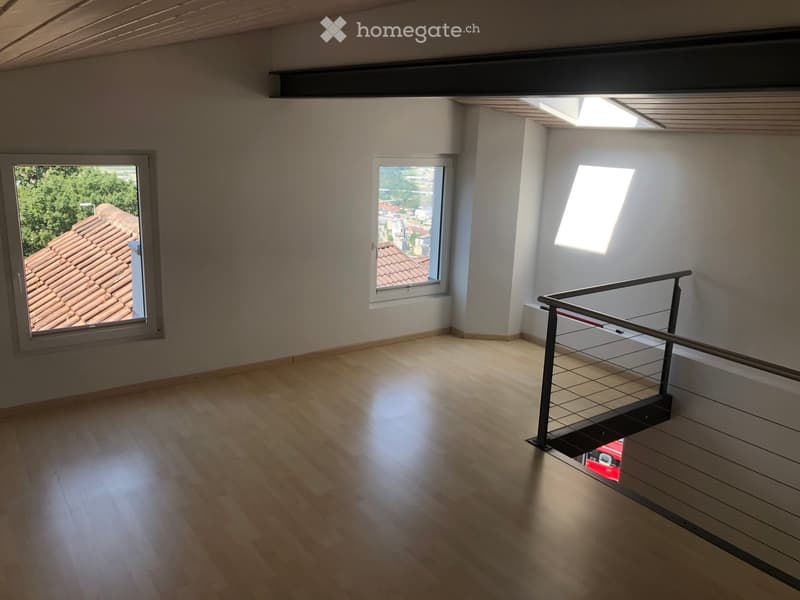 Empty room with wooden flooring, white walls, and large windows offering views of a tiled roof and surrounding area. A modern metal railing is visible, leading to an upper level.
