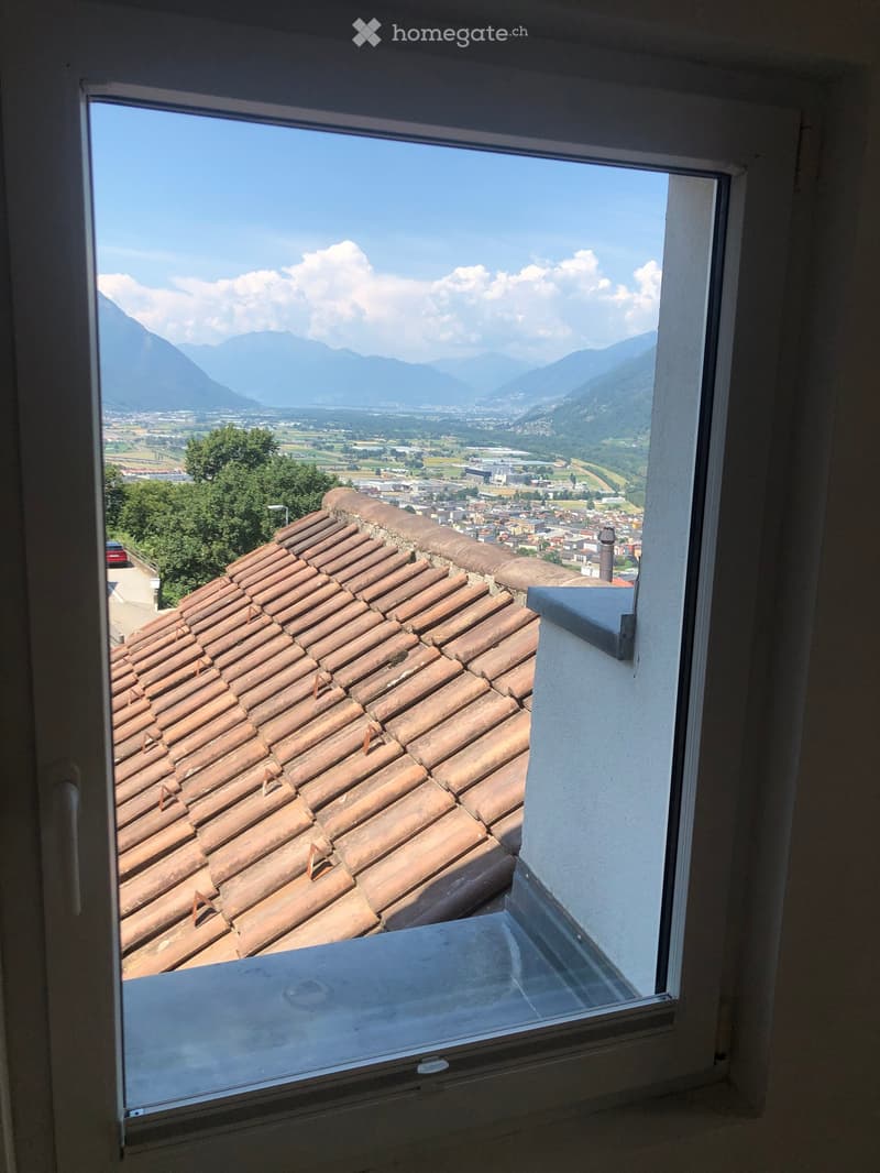 View from a balcony showing a terracotta-tiled roof, distant town, and mountain range under a clear blue sky.