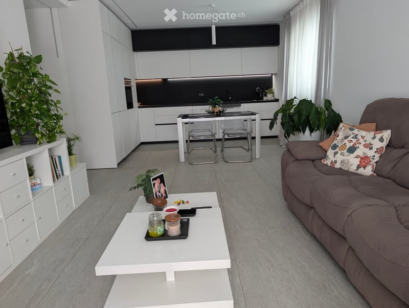 Modern living room with a grey sofa, white coffee table, dining area with chairs, and kitchen area with white cabinets and black countertops.