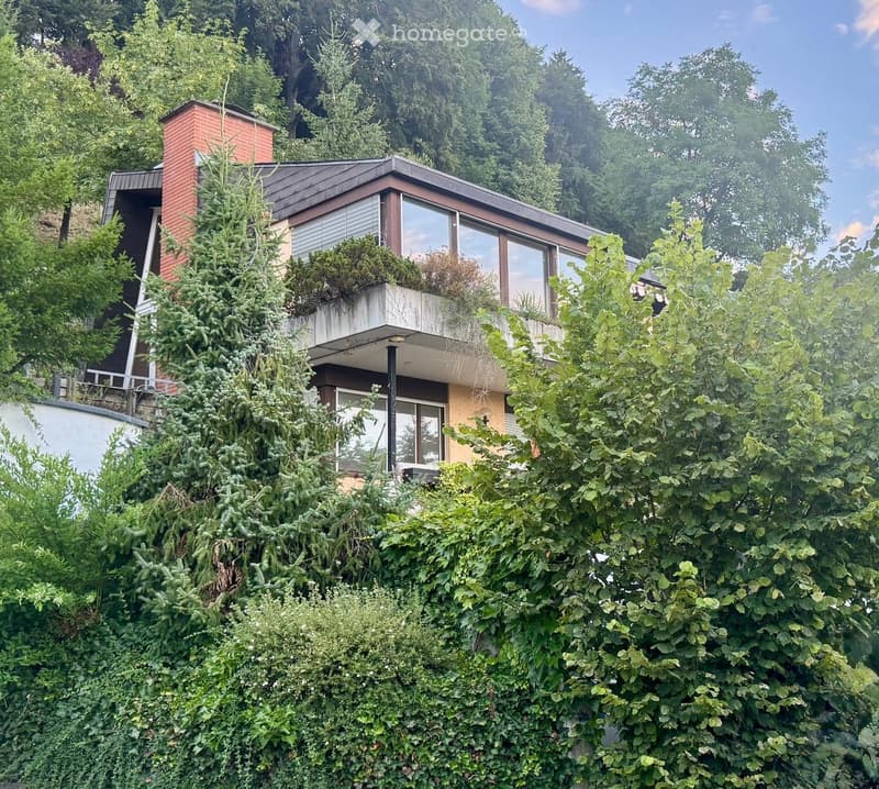 Modern two-story house with a flat roof, large windows, and a small balcony on the upper level, surrounded by lush greenery.