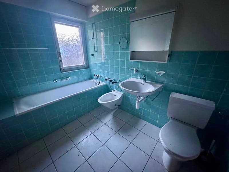 Bathroom with teal tiles, bathtub, toilet, sink, bidet, and a window with frosted glass.