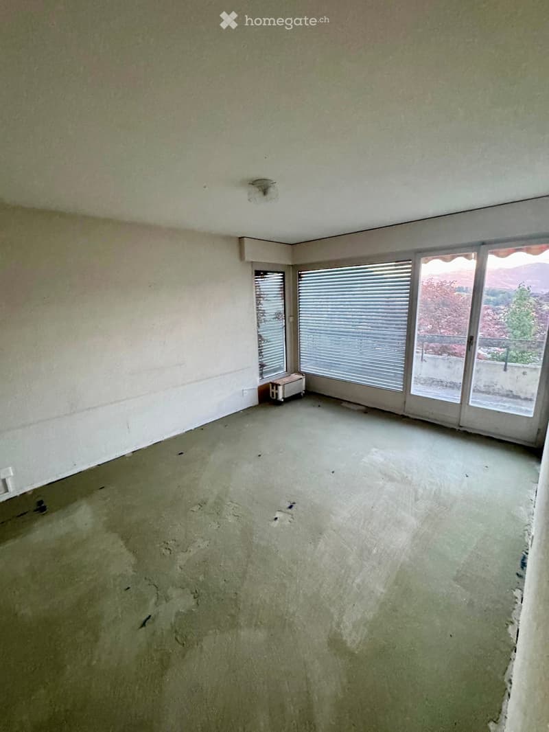 Empty room with unfinished concrete floor and walls, large windows with blinds, and a small radiator in the corner.