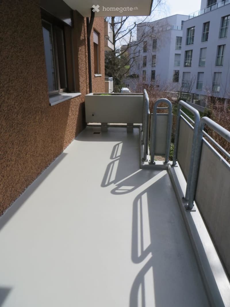 Clean, modern balcony with white flooring, metal railings, and a small planter box. The balcony is adjacent to a building with a textured brown wall and large windows.