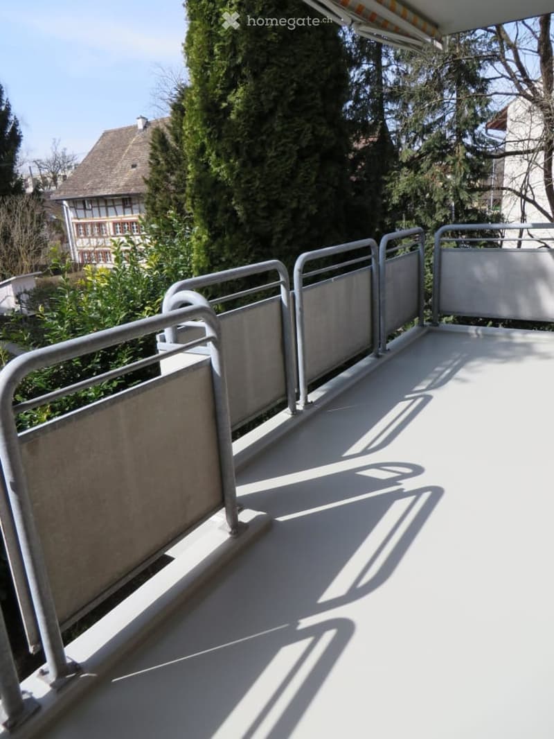 Balcony with metal railings, white floor, overlooking a garden and neighboring house.