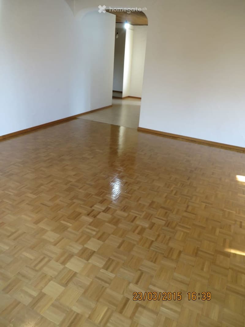 Empty room with polished wooden herringbone flooring and white walls, featuring a visible doorway leading to another space.