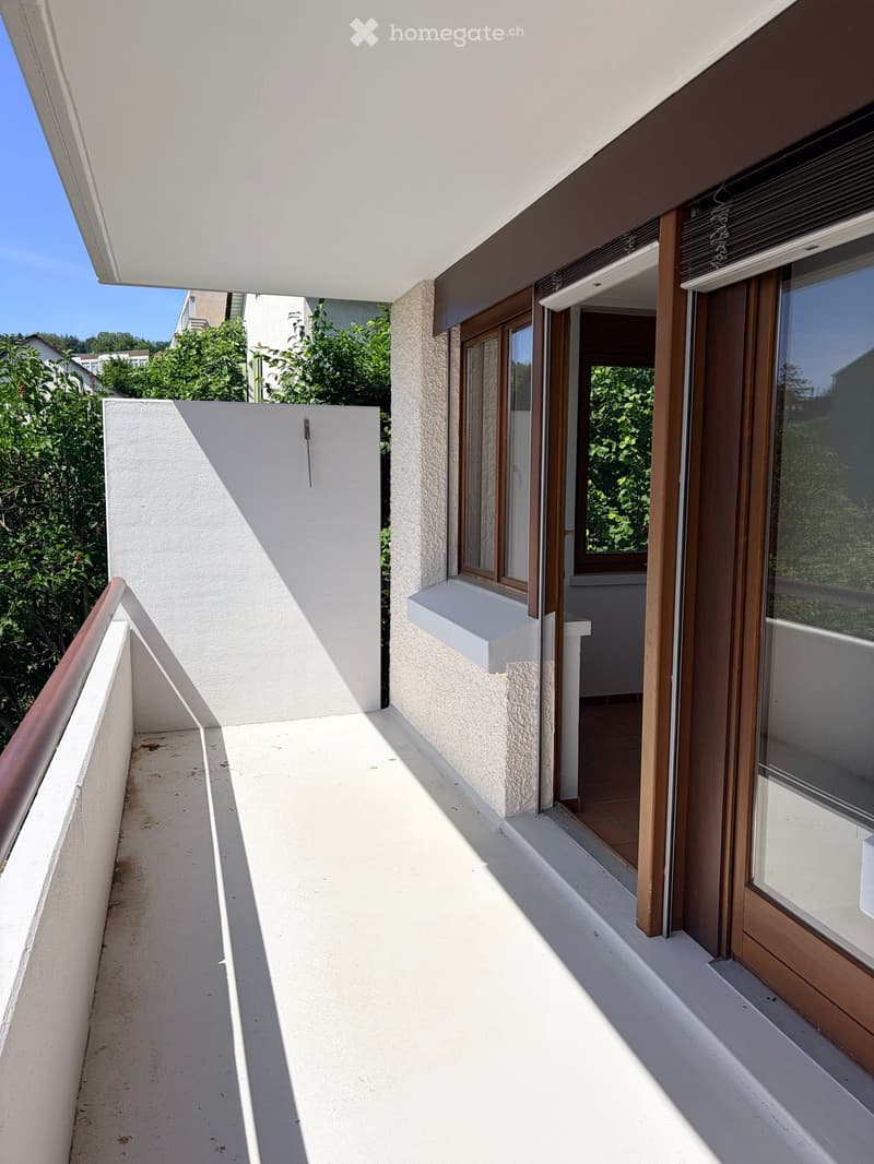 Balcony with white floor, wooden railings, and access to a room with tiled flooring and wooden window frames.