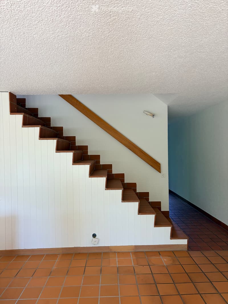 Staircase with wooden handrail and steps, white and orange tiled walls, and a light fixture on the wall.