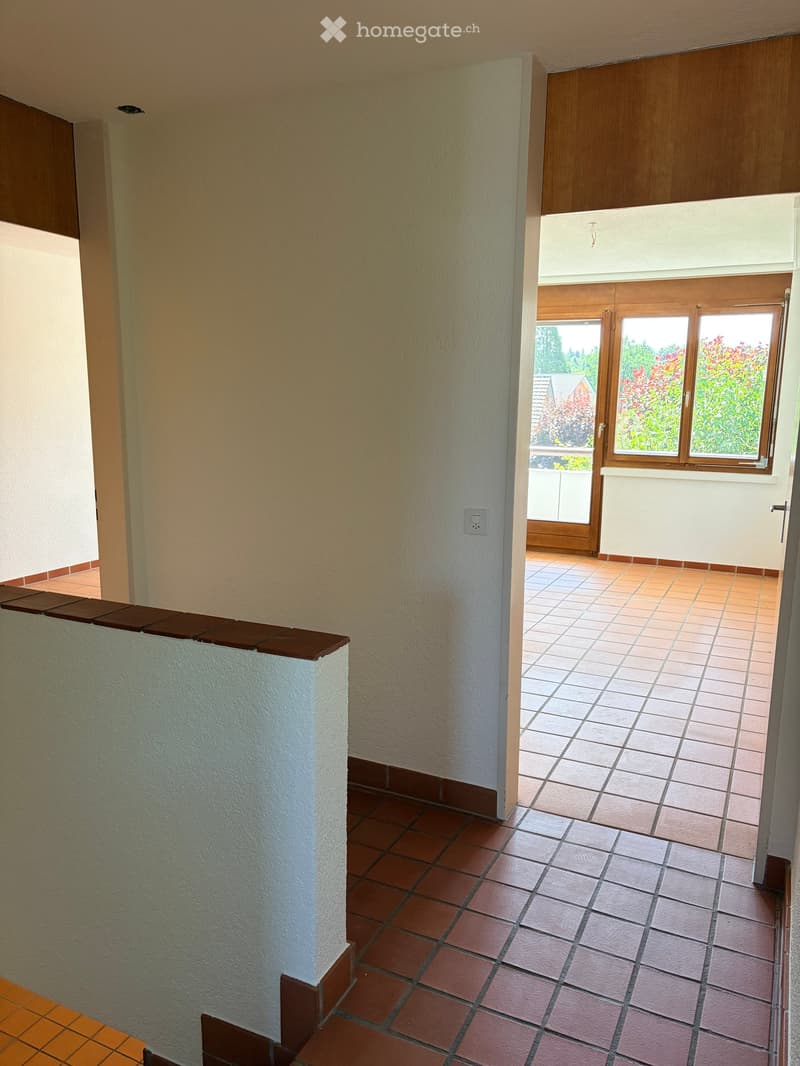 Hallway with tiled flooring, white walls, and a wooden door leading to a balcony area with outdoor tiles and a view of greenery.