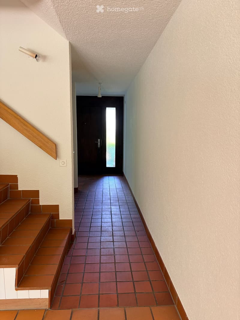 A narrow hallway with tiled flooring, beige walls, and a wooden staircase leading upwards. A door at the end provides access to another area.